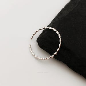 May include: A silver ring with a twisted rope design. The open-ended ring is set against a black and white background, highlighting its metallic sheen and unique texture. The ring's design is simple yet elegant, suitable for everyday wear.