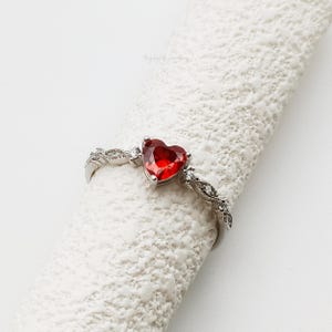 Puede incluir: Anillo de plata con una gema roja en forma de corazón. La banda está adornada con pequeñas piedras claras y un diseño retorcido. El anillo se muestra sobre una superficie blanca texturizada, resaltando su diseño.