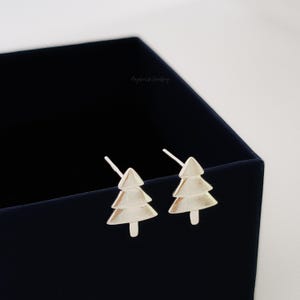 May include: A pair of silver-coloured stud earrings in the shape of Christmas trees. The earrings are presented on a dark blue surface. The earrings have a textured finish and a post backing.
