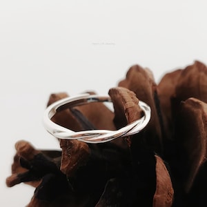 Puede incluir: Un anillo de plata pulida con un delicado diseño de banda trenzada. El anillo tiene una superficie metálica lisa y reflectante con un patrón entrelazado continuo que crea un aspecto sutil y elegante. La banda es circular y uniforme.