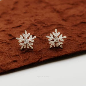 May include: A pair of silver snowflake stud earrings. Each earring has a detailed, symmetrical design with six points, and a small, clear gemstone at the centre. The earrings are displayed on a brown surface.