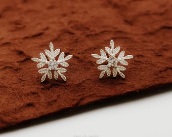 May include: A pair of silver stud earrings shaped like intricate snowflakes. Each earring features a central clear gemstone surrounded by delicate, textured metallic branches that radiate outward in a symmetrical, multi-pointed star pattern.