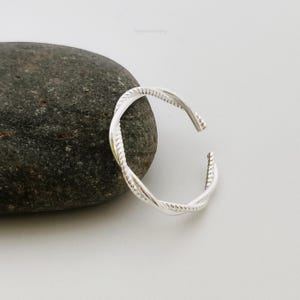 May include: A silver twisted ring with a rope-like design. The ring is on a grey rock.