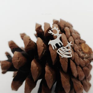 May include: A pair of silver-toned stud earrings featuring a reindeer silhouette and a stylised Christmas tree shape. The earrings have a matte finish and are displayed against a brown pinecone, showcasing a festive theme with clean lines.