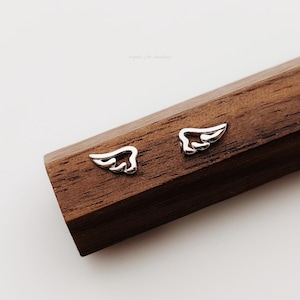 Puede incluir: Dos pequeños pendientes de botón plateados con forma de ala. Los pendientes se muestran sobre una superficie de madera marrón oscuro. Las alas tienen un diseño detallado y con plumas.