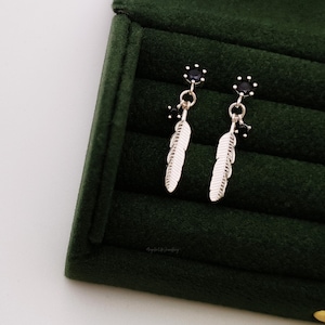 Puede incluir: Un par de pendientes de plumas plateadas con detalles de piedras preciosas negras. Los pendientes se muestran sobre un fondo de terciopelo verde.