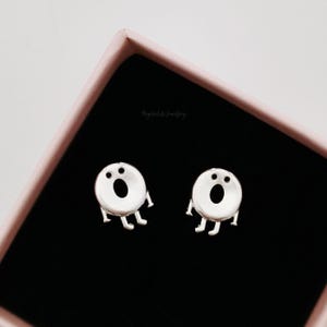 Puede incluir: Un par de pendientes de botón de plata con forma de cara de dibujos animados con dos ojos y una boca. Los pendientes están en una pequeña caja rosa.