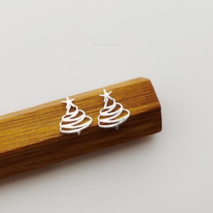 May include: A pair of silver-toned stud earrings shaped like stylised Christmas trees with a textured finish. Each earring features a pointed star at the top and three horizontal, curved lines forming the tree body, resting on a wooden block.