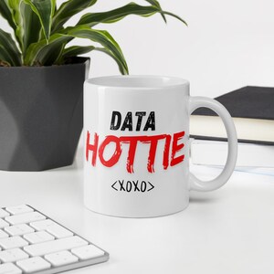 May include: White ceramic mug with the words "DATA HOTTIE" in black and red, with "<XOXO>" below. The mug is on a white surface with a keyboard, a plant, and a book in the background.