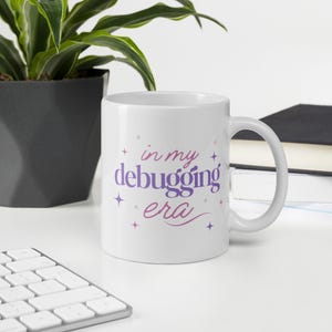 May include: White ceramic mug with the text "in my debugging era" in purple and pink script. The mug has a curved handle and is surrounded by a plant in a dark gray pot, a keyboard, and a stack of books.