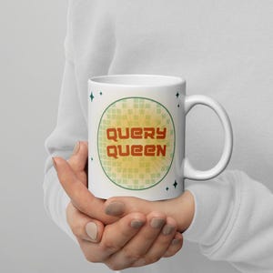 Op de afbeelding: Witte keramische mok met de tekst "QUERY QUEEN" in een retro lettertype, binnen een groen omcirkelde cirkel met een geel en groen geometrisch patroon. De mok wordt met beide handen vastgehouden.