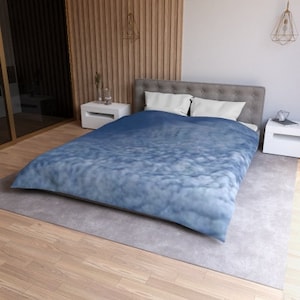 Pode incluir: Uma cama com uma colcha com padrão de nuvens azuis e brancas. A cama tem almofadas brancas e está sobre um tapete cinza. O quarto tem chão de madeira e uma parede de destaque em madeira.