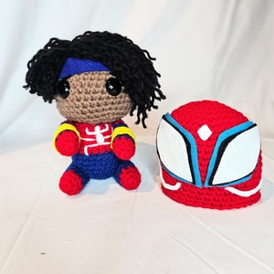 May include: Two crocheted plush toys. One is a red and blue superhero mask with white accents. The other is a brown-skinned person with black hair wearing a red, blue, and yellow costume.
