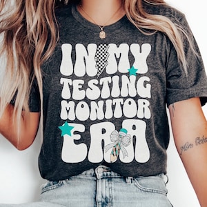 May include: A dark grey heather t-shirt with the text "In My Testing Monitor Era" in white with a blue and white lightning bolt and a blue bow with a star on top.