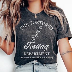 May include: A dark grey heather t-shirt with a white graphic of a pencil and the text "The Tortured Testing Department. All's fair in academic monitoring."