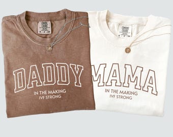 May include: Two t-shirts, one brown and one cream, with the words "DADDY" and "MAMA" printed in white block letters. The text "IN THE MAKING IVF STRONG" is below. Each shirt has a silver necklace.