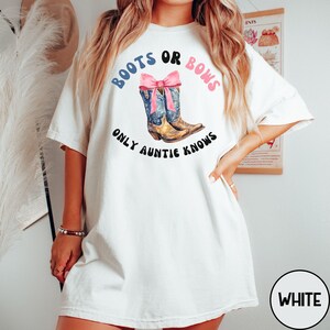 May include: A white short-sleeve t-shirt featuring a graphic of two brown and blue cowboy boots tied together with a pink bow. The text reads "BOOTS OR BOWS" in blue and pink, and "ONLY AUNTIE KNOWS" in black, arranged in a circular pattern.