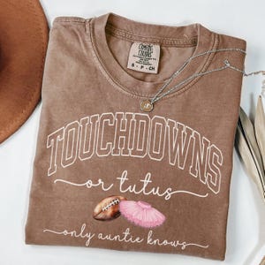 May include: A brown Comfort Colors t-shirt with the words "TOUCHDOWNS or tutus" in white script, along with a football and pink tutu graphic. A silver necklace with a pendant is draped across the shirt.