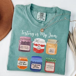May include: A teal Comfort Colors t-shirt with the phrase "Testing is My Jam" and illustrations of six jam jars, each with an encouraging message. A silver necklace with a pendant is draped across the shirt.