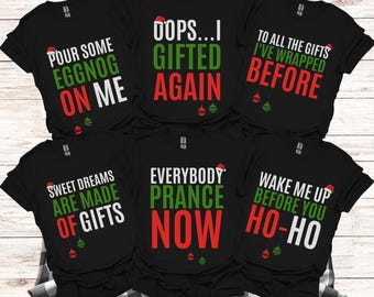 May include: Six black short-sleeved t-shirts featuring Christmas-themed text and graphics. Each shirt displays unique holiday phrases in white, green, and red lettering, accented with small Santa hats and hanging ornament illustrations.