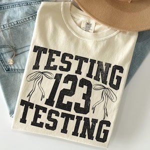 May include: Cream-colored t-shirt with the words "TESTING 123 TESTING" in distressed black lettering. The design includes bow illustrations. The shirt is laid on top of blue jeans and a tan hat.