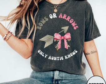 Könnte beinhalten: Ein anthrazitfarbenes Kurzarm-T-Shirt mit einem Grafikdruck und dem Text "Bows or Arrows Only Auntie Knows". Das Design zeigt eine hellrosa Schleife und einen Pfeil mit Tarnmuster. Das T-Shirt hat einen Rundhalsausschnitt und eine lockere Passform.