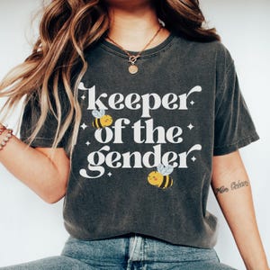 May include: Dark gray t-shirt with the text "keeper of the gender" in white script. The shirt features two cartoon bees with yellow and black stripes. The shirt is a casual style.