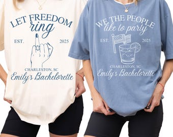 May include: Two t-shirts, one cream and one blue, with graphic text. The cream shirt says "LET FREEDOM ring" with a hand gesture and "Emily's Bachelorette" below. The blue shirt says "WE THE PEOPLE like to party" with a cocktail graphic and "Emily's Bachelorette".