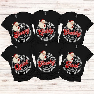 May include: Six black t-shirts with a white circle design featuring a cartoon Santa Claus and text. The text on the shirts reads "The Sleepy Shopper", "The Giddy Shopper", "The Crabby Shopper", "The Quiet Shopper", "The Flashy Shopper", and "The Fast Shopper".