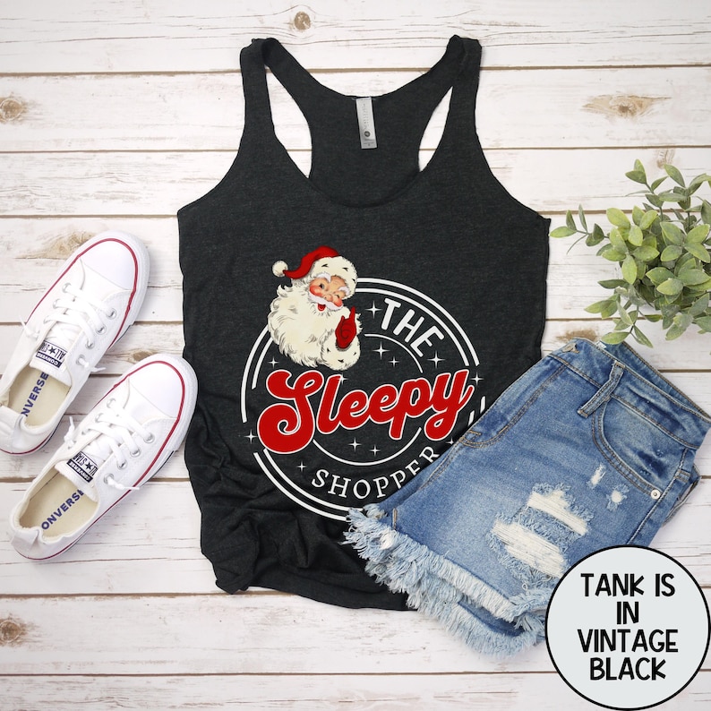 May include: A black tank top with a white circle graphic featuring a cartoon Santa Claus and the text "The Sleepy Shopper".