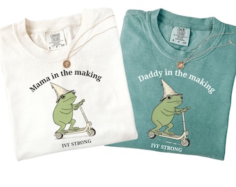 May include: Two Comfort Colors t-shirts, one cream and one teal, with matching gold necklaces. The cream shirt says "Mama in the making" and the teal shirt says "Daddy in the making." Both shirts feature a green frog riding a scooter and the text "IVF STRONG."
