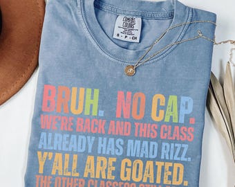 May include: A blue Comfort Colors t-shirt with the text "BRUH. NO CAP. WE'RE BACK AND THIS CLASS ALREADY HAS MAD RIZZ. Y'ALL ARE GOATED. THE OTHER CLASSES? STILL MID." in colorful letters. A gold necklace is draped over the shirt.
