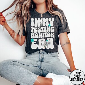 May include: A dark grey heather t-shirt featuring white retro-style text that reads IN MY TESTING MONITOR ERA. The design includes a black and white checkered lightning bolt, two teal stars, and a pencil graphic adorned with a leopard print bow.