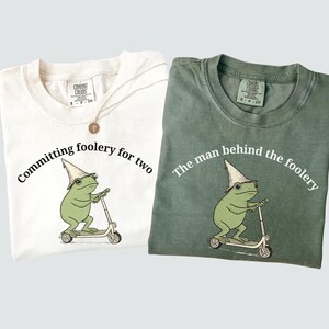 May include: Two t-shirts, one cream and one green, with a frog graphic. The cream shirt says "Committing foolery for two." The green shirt says "The man behind the foolery." Both shirts feature a frog riding a scooter.