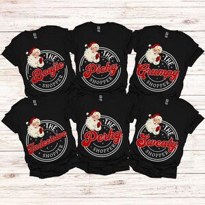 May include: Six black t-shirts with a white circle design featuring a cartoon Santa Claus and text that reads "The [adjective] Shopper". The adjectives are "Bougie", "Picky", "Grumpy", "Indecisive", "Perky", and "Sweaty".