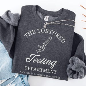 May include: A dark gray sweatshirt with the text "THE TORTURED TESTING DEPARTMENT" in white, along with a pencil graphic. The phrase "All's fair in academic monitoring" is below. A silver necklace with a sun pendant is draped across the sweatshirt.