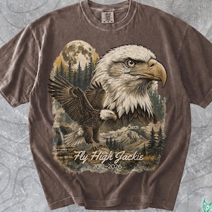 May include: A brown short-sleeve t-shirt featuring a graphic print of a bald eagle head and a flying eagle over a forest and mountain landscape with a full moon. The text reads Fly High Jackie 2012-2026 in a white script font below the eagle.