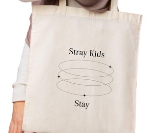 May include: Cream-colored tote bag with the text "Stray Kids" above a design of three swirling lines and the word "Stay" below. The bag is held by a person wearing a white long-sleeve shirt, a brown scarf, and pink pants.