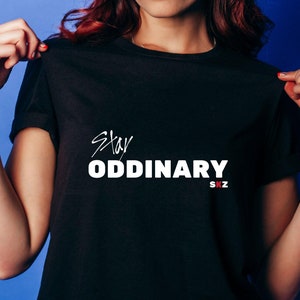 May include: Black t-shirt featuring the words "Stay Oddinary" in white script and block letters. The letters "SKZ" are in red below the word "Oddinary". The shirt is being held up by a person.