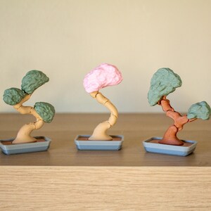May include: Three miniature bonsai trees in different colors. The trees are made of plastic and have a realistic look. The trees are in gray pots with brown bases.