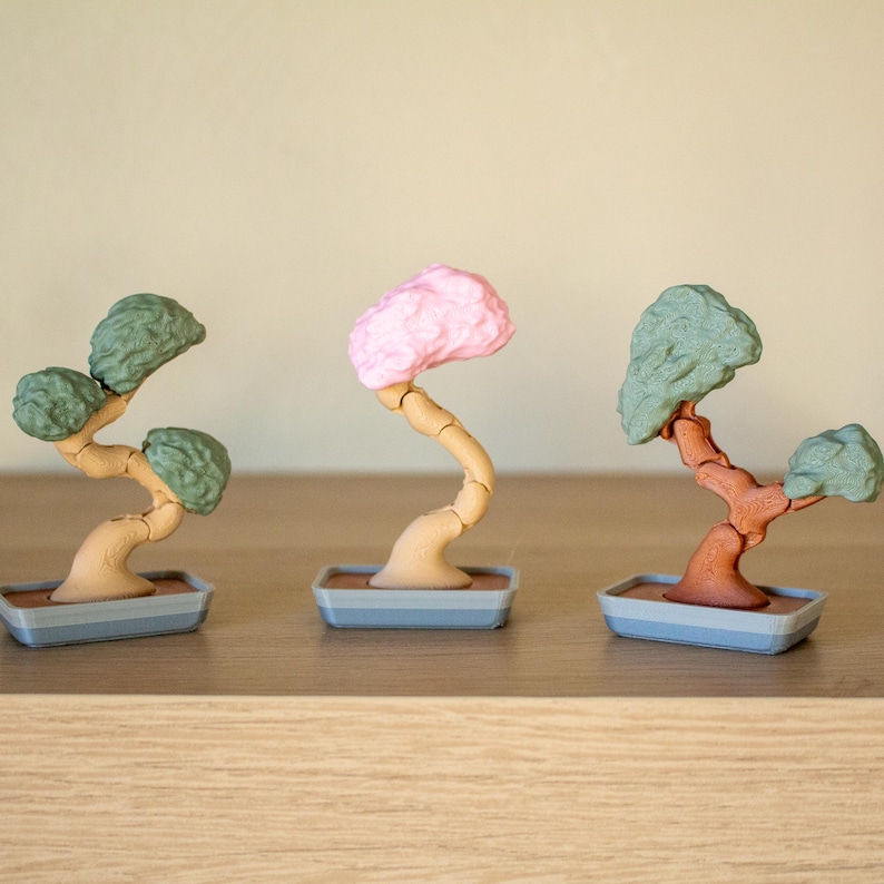 May include: Three miniature bonsai trees in different colors. The trees are made of plastic and have a realistic look. The trees are in gray pots with brown bases.
