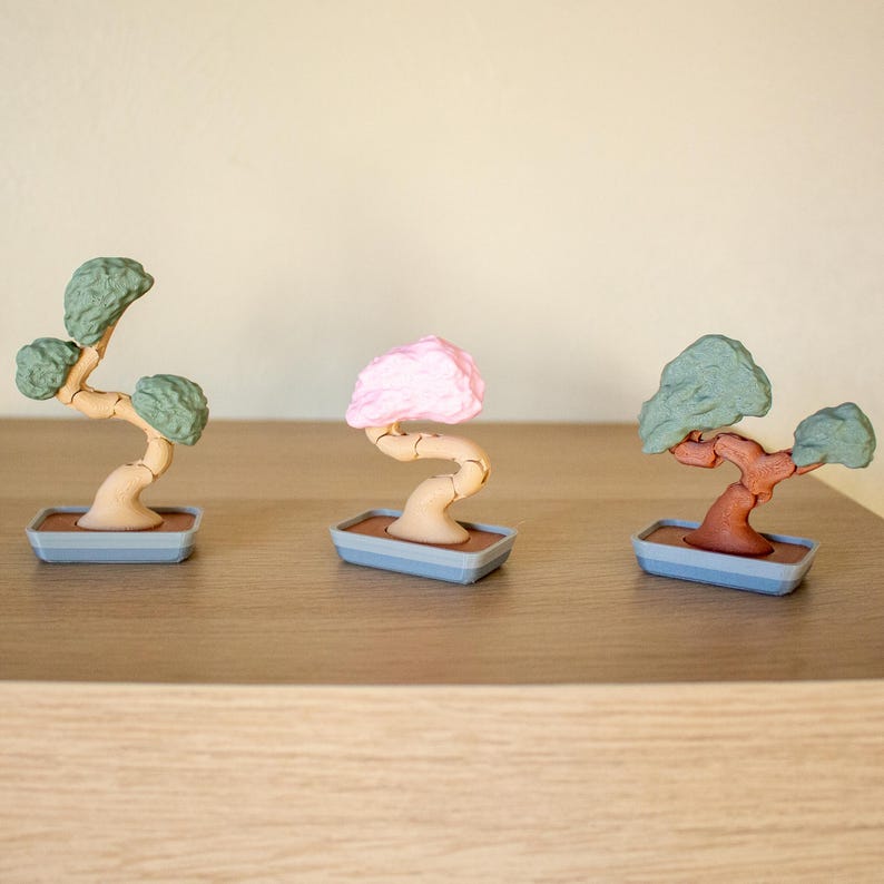 May include: Three miniature bonsai trees in pots. The trees are made of plastic and have different colored leaves: green, pink, and brown. The pots are gray with a brown base.