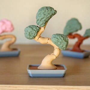 May include: A set of three miniature bonsai trees, each with a different colored base and foliage. The trees are made of plastic and have a realistic appearance. The trees are arranged on a wooden surface.