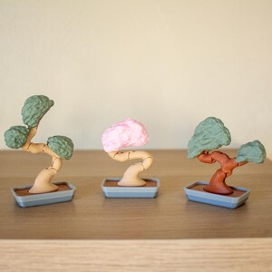 May include: Three miniature bonsai trees in different colors. The trees are made of plastic and have a brown trunk and green, pink, and brown leaves. The trees are sitting on small gray pots.