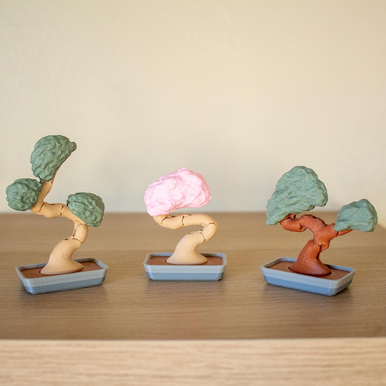 May include: Three miniature bonsai trees in different colors. The trees are made of plastic and have a brown trunk and green, pink, and brown leaves. The trees are sitting on small gray pots.