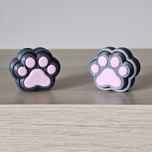 May include: Two paw-shaped decorative objects. Each paw has a dark gray outer layer and a light pink paw print in the center. The objects are sitting on a light brown surface.