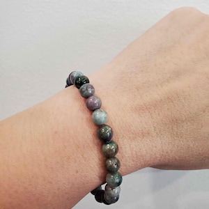 May include: A stretch bracelet composed of smooth, round beads in varying shades of dark green, deep purple, and muted blue. The beads are strung together in a single strand, creating a multi-coloured, polished appearance on the wrist.