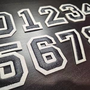 May include: Close-up of embroidered black numbers 0 to 9 with white borders. The numbers are in a collegiate style, with a thick, bold font. The patches are likely for sports jerseys or other apparel.