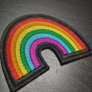 May include: Embroidered rainbow patch with a vibrant spectrum of colours: red, orange, yellow, green, blue, and purple. The rainbow is outlined with a black border. Suitable for clothing, bags, or other fabric items.