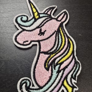 May include: Embroidered unicorn patch. The design features a pink body, a yellow horn, and a flowing mane in pastel shades. The patch has a white border and is suitable for application on clothing or accessories.